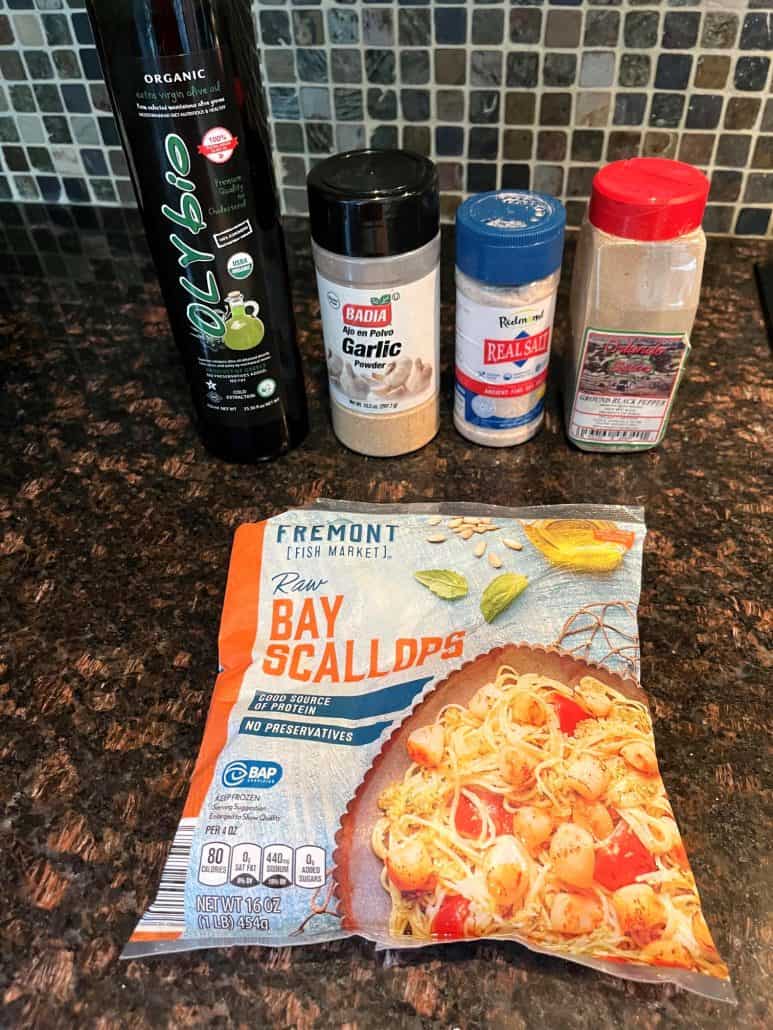 Ingredients for Pan Fried Bay Scallops recipe, including a package of raw bay scallops, olive oil, garlic powder, sea salt, and black pepper, arranged on a kitchen counter.