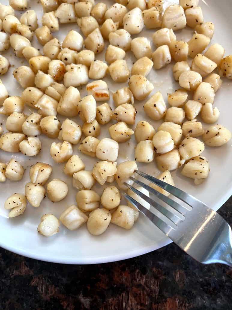 Cooked bay scallops on a white plate, with a fork touching one scallop.