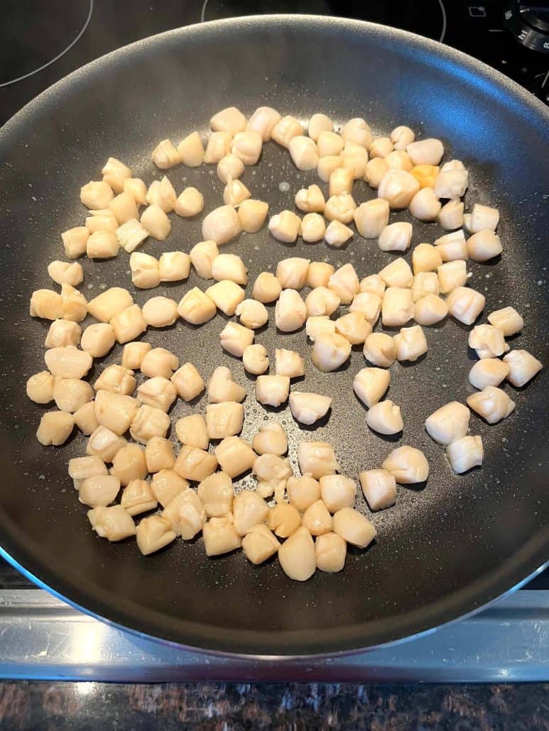 Bay scallops cooking in a non-stick pan, beginning to turn opaque as they sizzle.