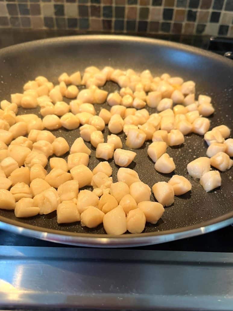 Bay scallops cooking in a non-stick pan, starting to turn golden as they sizzle, part of the Pan Fried Bay Scallops recipe process.