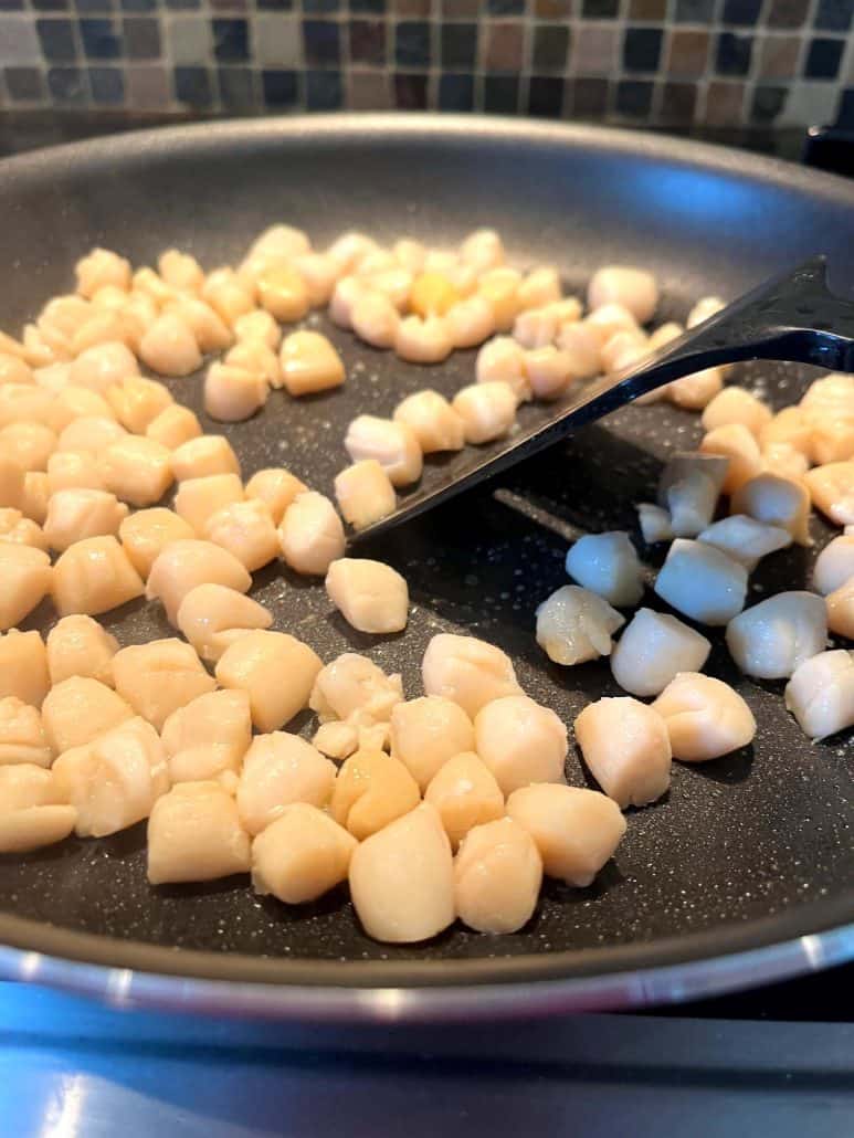 Close-up of bay scallops being cooked in a non-stick pan with a spatula stirring them.