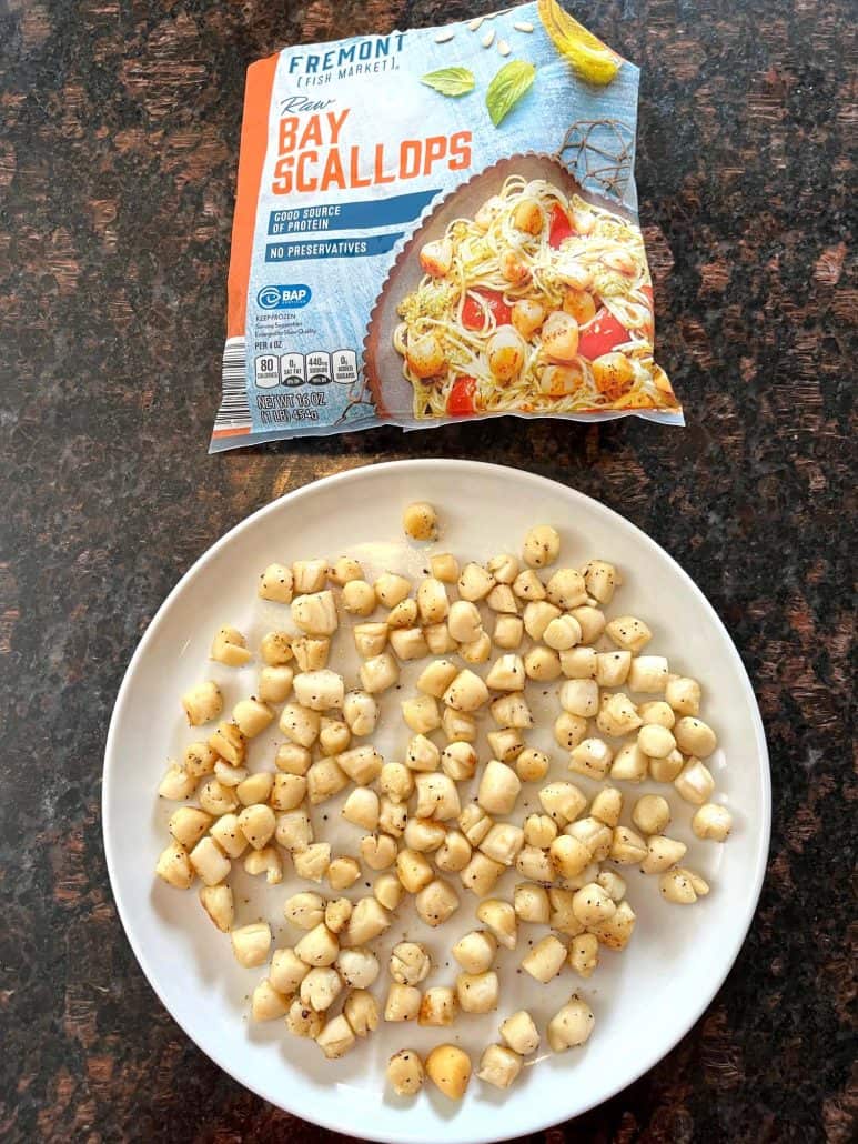 Cooked bay scallops served on a white plate, with a package of raw bay scallops in the background, showcasing the final result of the Pan Fried Bay Scallops recipe.