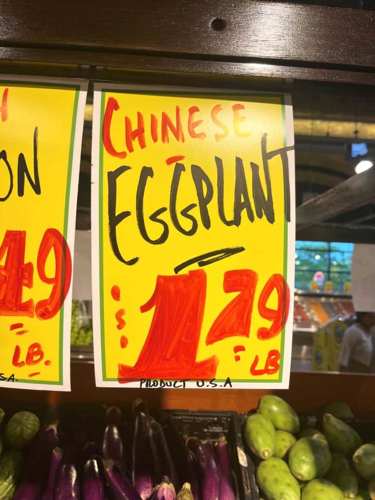 A brightly colored sign at a grocery store advertising Chinese eggplant for $1.79 per pound, with fresh Chinese eggplants displayed below.