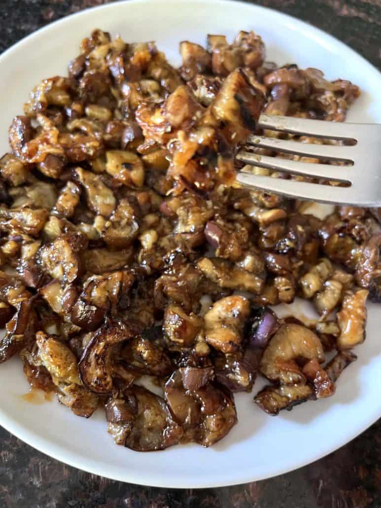 A fork holding a bite of pan-fried Chinese eggplant with garlic sauce above a plate, showcasing the tender, caramelized eggplant pieces coated in a flavorful sauce.