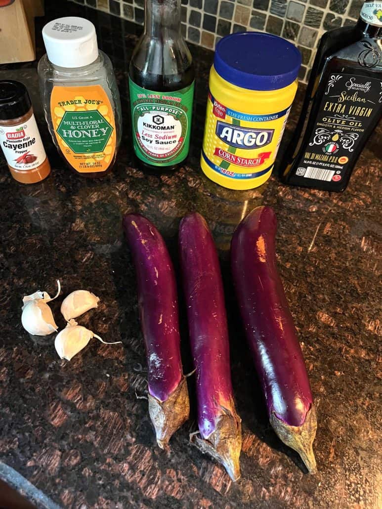 Ingredients for pan-fried Chinese eggplant with garlic sauce, including fresh Chinese eggplants, garlic cloves, cayenne pepper, honey, reduced-sodium soy sauce, cornstarch, and extra virgin olive oil, laid out on a kitchen countertop, ready for cooking.