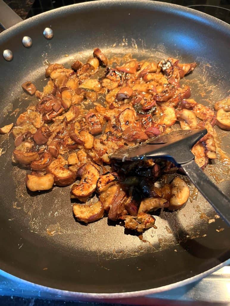 A skillet filled with pan-fried Chinese eggplant in garlic sauce, with a spatula stirring the caramelized eggplant pieces to ensure even cooking and coating.