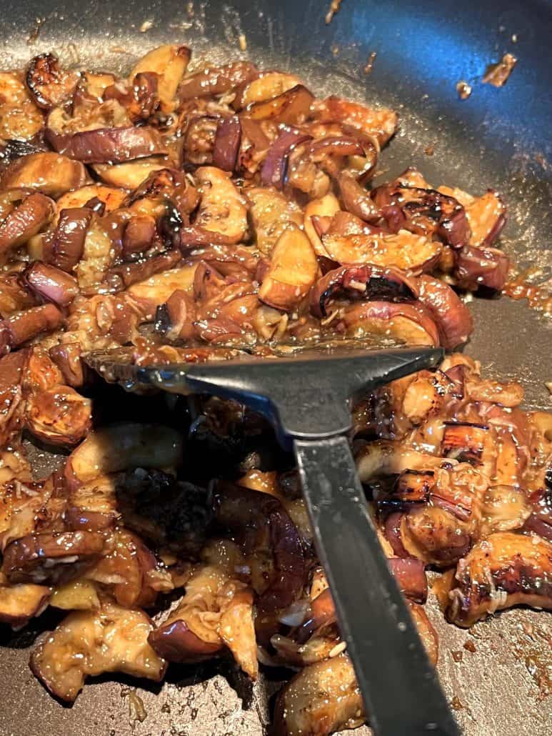 Pan-fried Chinese eggplant pieces cooking in a skillet with a rich garlic sauce, being stirred with a spatula to ensure even coating and cooking.