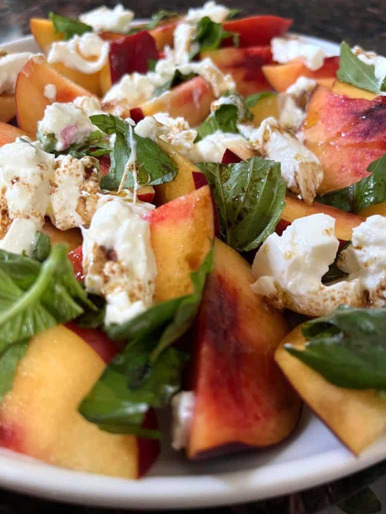 Whole peach burrata salad seen on a white plate from a close angle.