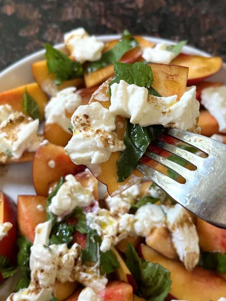 Fork piercing a fresh peach burrata salad with fresh peaches, creamy burrata, and basil leaves.