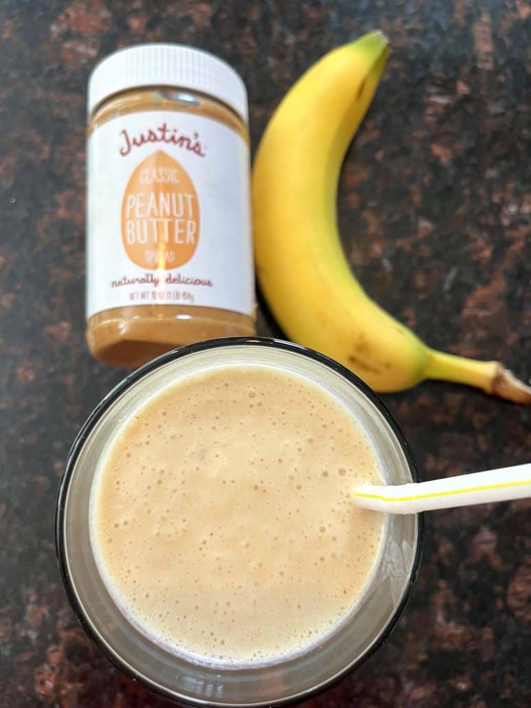 Top view of Peanut Butter Banana Smoothie with a yellow straw, alongside a jar of Justin's peanut butter and a ripe banana