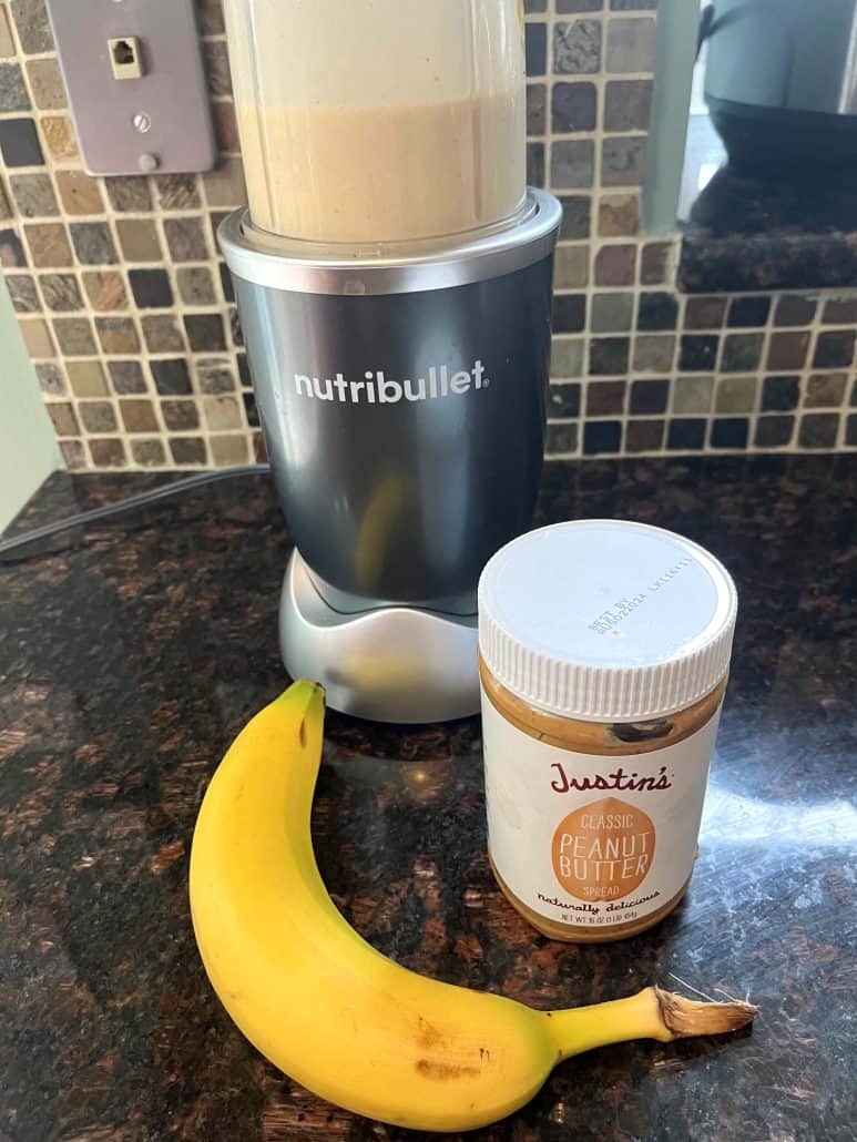 A Nutribullet blender blending with banana and a jar of Justin's peanut butter placed in front on the kitchen countertop.