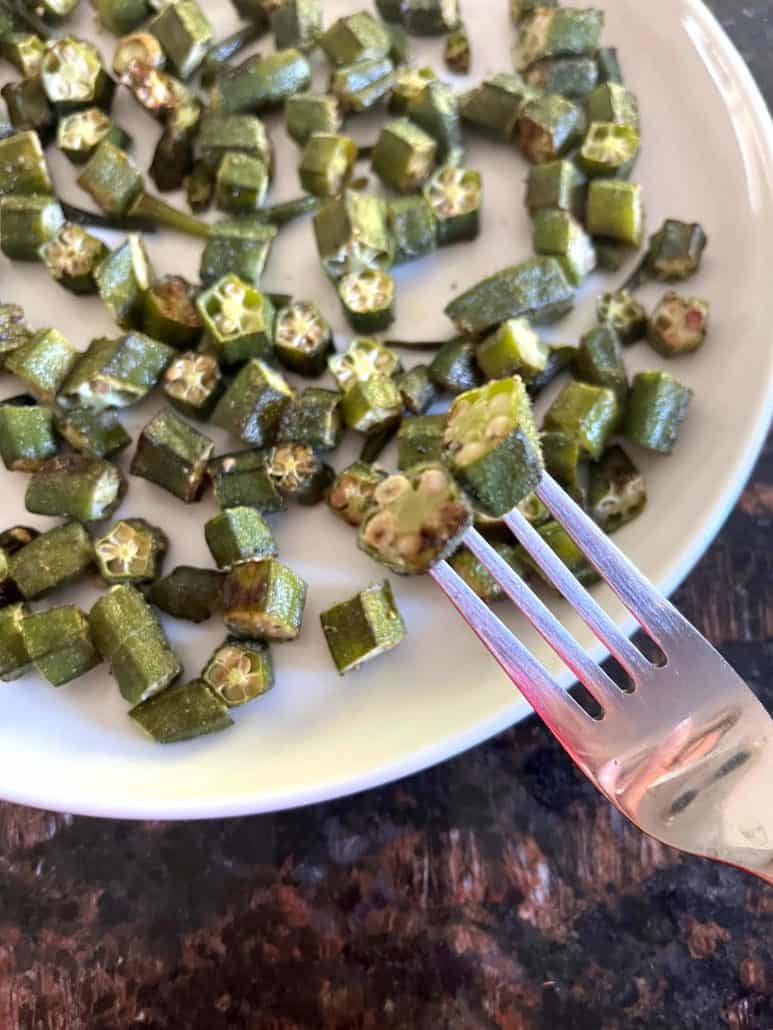 A fork holding a piece of roasted okra above a white plate filled with crispy roasted okra pieces.