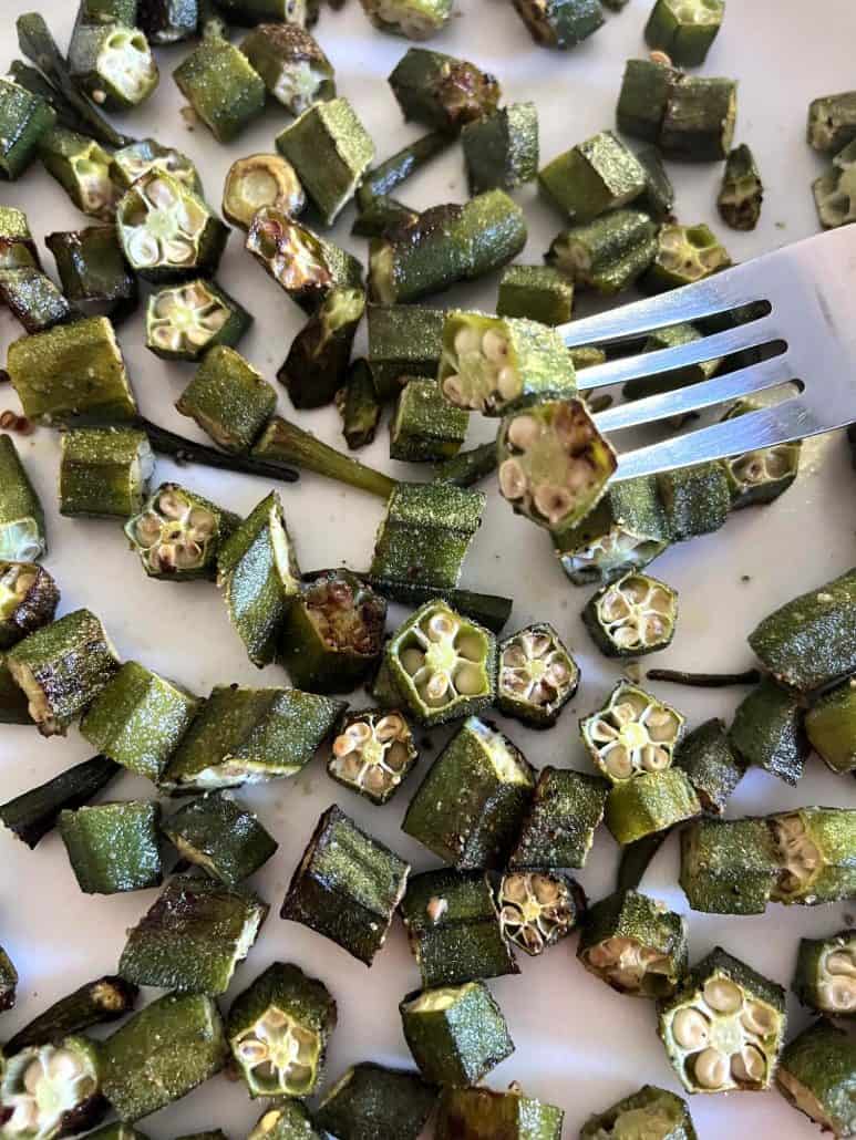 Close-up of a fork holding a roasted okra slice above a plate filled with crispy roasted okra.