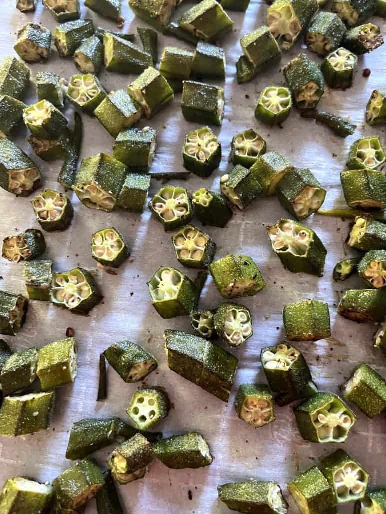 Sliced okra roasting on a parchment-lined baking sheet, evenly spaced to achieve crispy, golden brown edges.