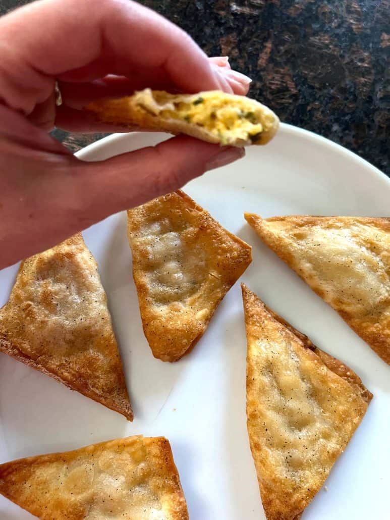 Hand of Melanie Mendelson from melaniecooks.com holding a Buffalo Style Chicken Popper with a bite taken, showing the creamy chicken filling, with more poppers on a plate below.