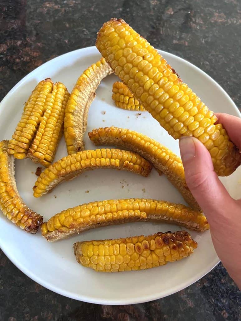 Melanie Mendelson from melaniecooks.com holding a crispy, air-fried corn rib from a plate of seasoned corn ribs, ready to eat.