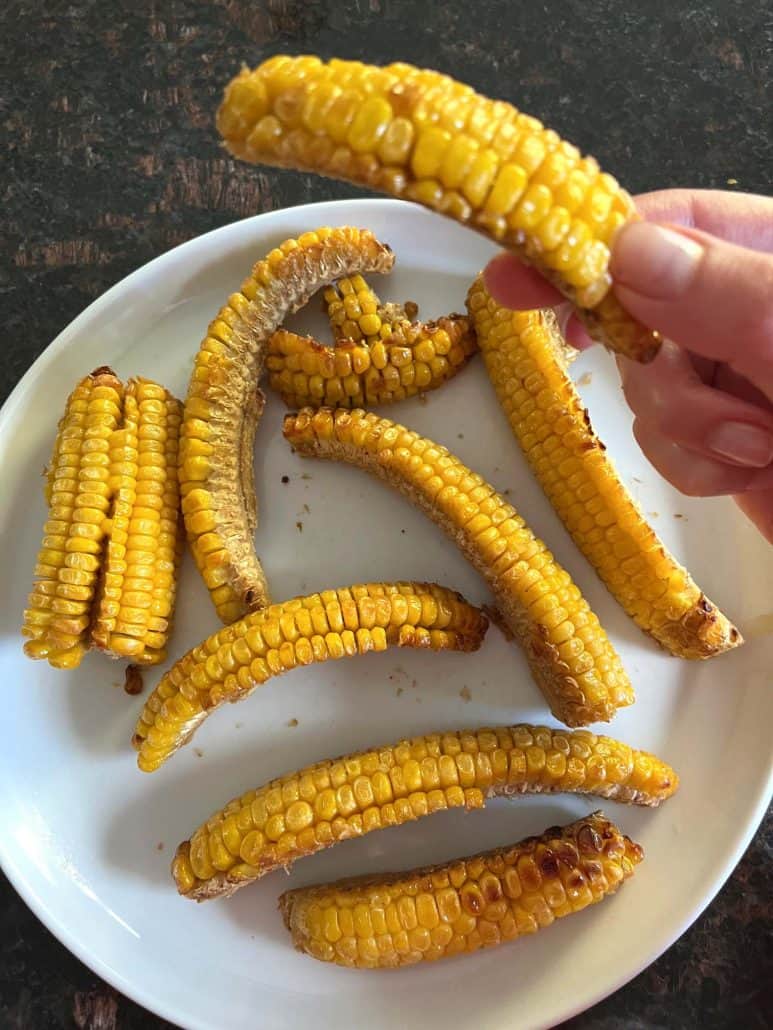 Melanie Mendelson from melaniecooks.com holding a seasoned, crispy corn rib from a plate of freshly air-fried corn ribs