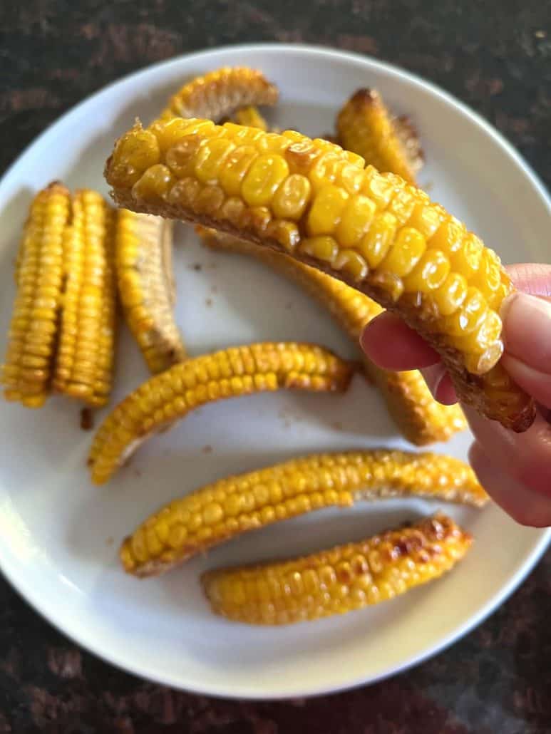 Close-up of a golden, air-fried corn rib held above a plate of seasoned corn rib.