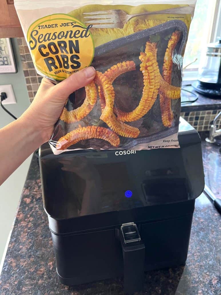 Melanie Mendelson from melaniecooks.com holding a package of Trader Joe's Seasoned Corn Ribs above an air fryer, ready for cooking straight from frozen.