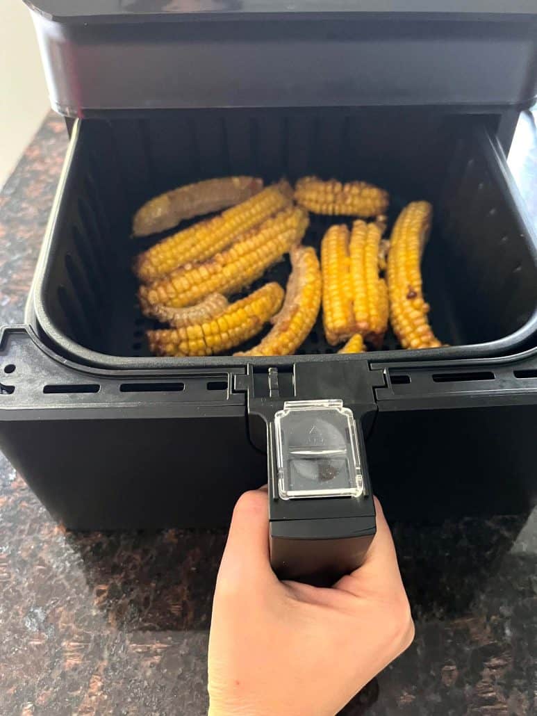 Hand of Melanie Mendelson from melaniecooks.com pulling out the air fryer basket with freshly cooked, golden-brown seasoned corn ribs.