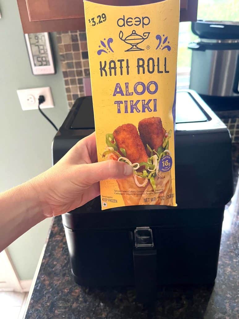 Holding a package of frozen Kati Roll Aloo Tikki in front of an air fryer.