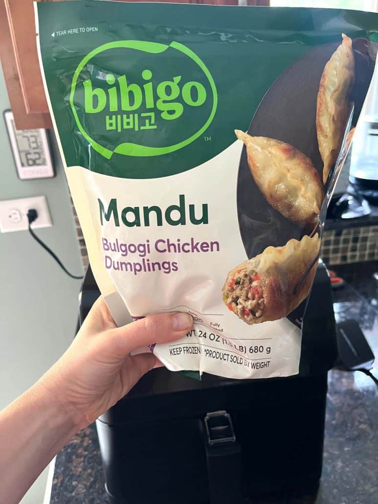 Hand holding a bag of Bibigo Mandu Korean Chicken Dumplings in front of an air fryer.