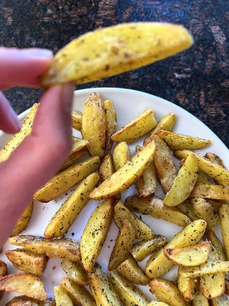 Hand of Melanie Mendelson from melaniecooks.com holding a crispy air-fried potato wedge above a plate.