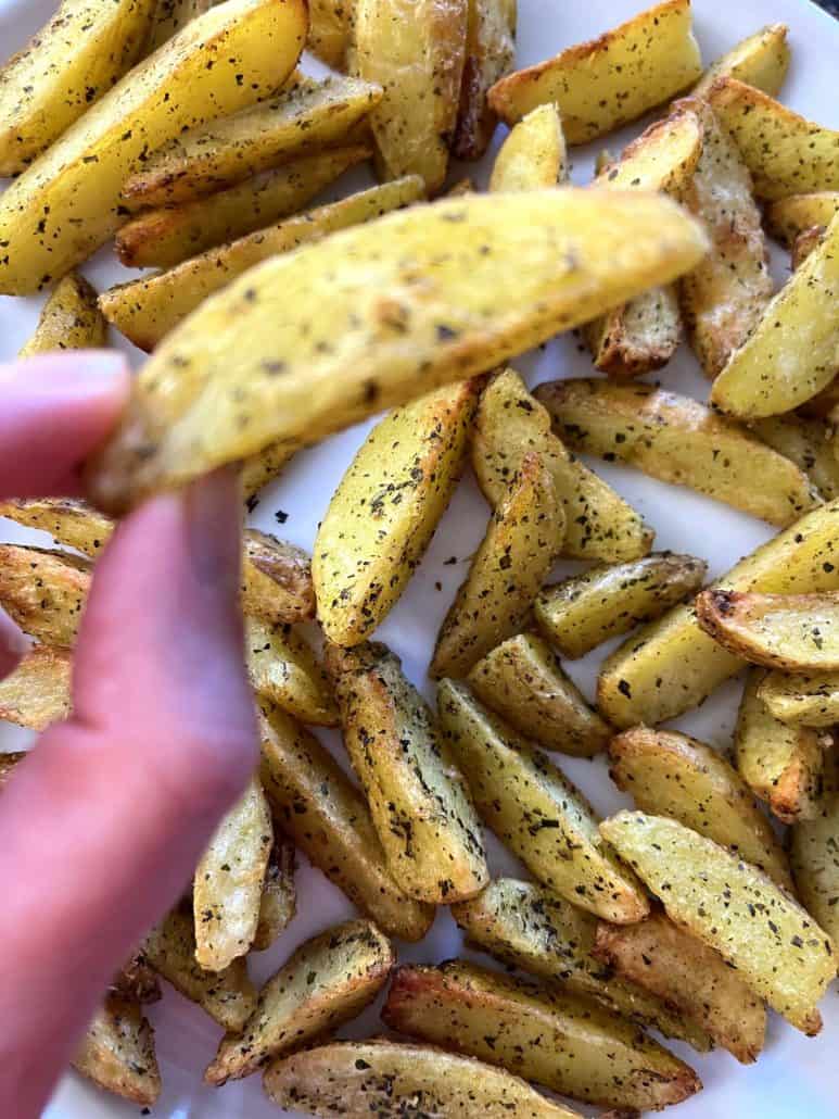 Hand of Melanie Mendelson from melaniecooks.com holding a perfectly crispy, golden air-fried potato wedge over a plate full of seasoned wedges.