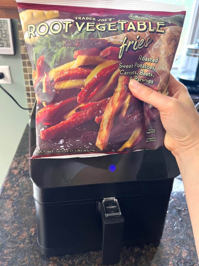 Hand of Melanie Mendelson from melaniecooks.com holding a bag of Trader Joe's Root Vegetable Fries above an air fryer, preparing to cook a healthy and colorful snack.