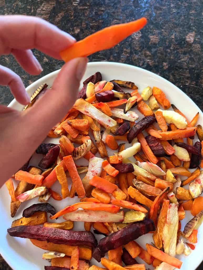 Melanie Mendelson from melaniecooks.com holding a crispy root vegetable fry above a plate of freshly air-fried root vegetable fries, showcasing their vibrant colors and texture.