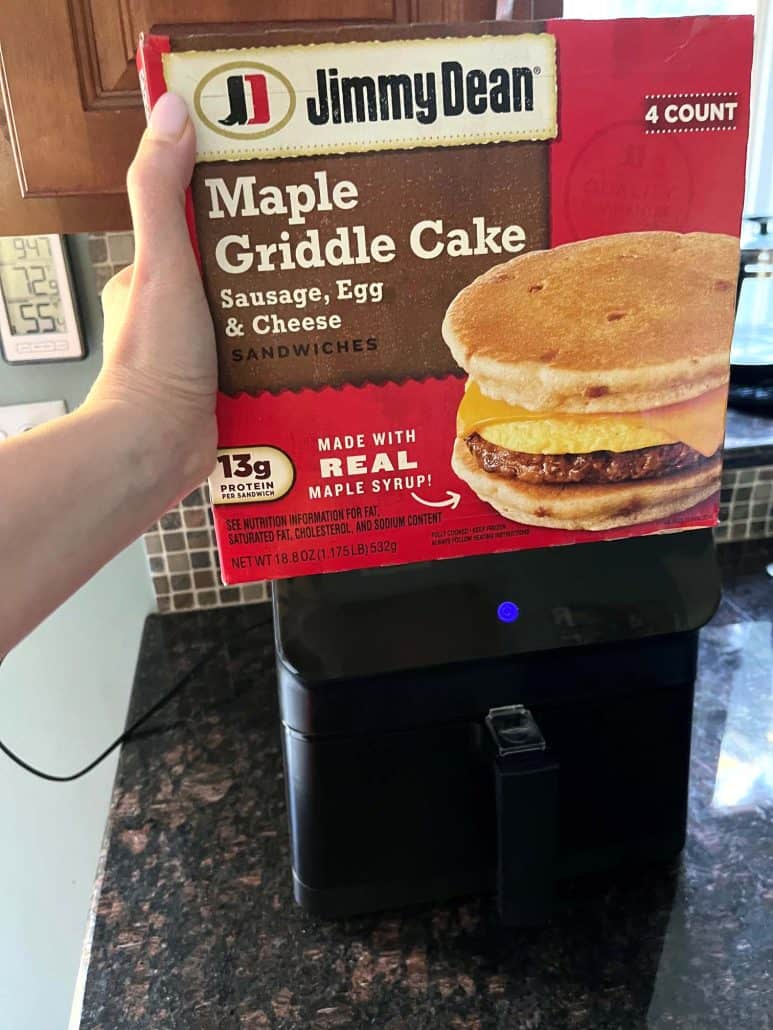 Holding a box of Jimmy Dean Maple Griddle Cake Sausage, Egg & Cheese Sandwiches above an air fryer, preparing to cook the sandwiches following an air fryer recipe.