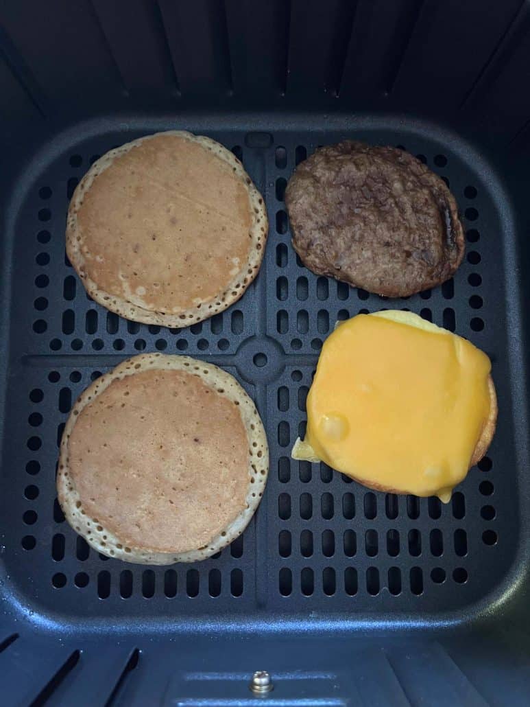 Close-up of air fryer basket, including pancakes, sausage patty, and egg with melted cheese, ready for assembly.