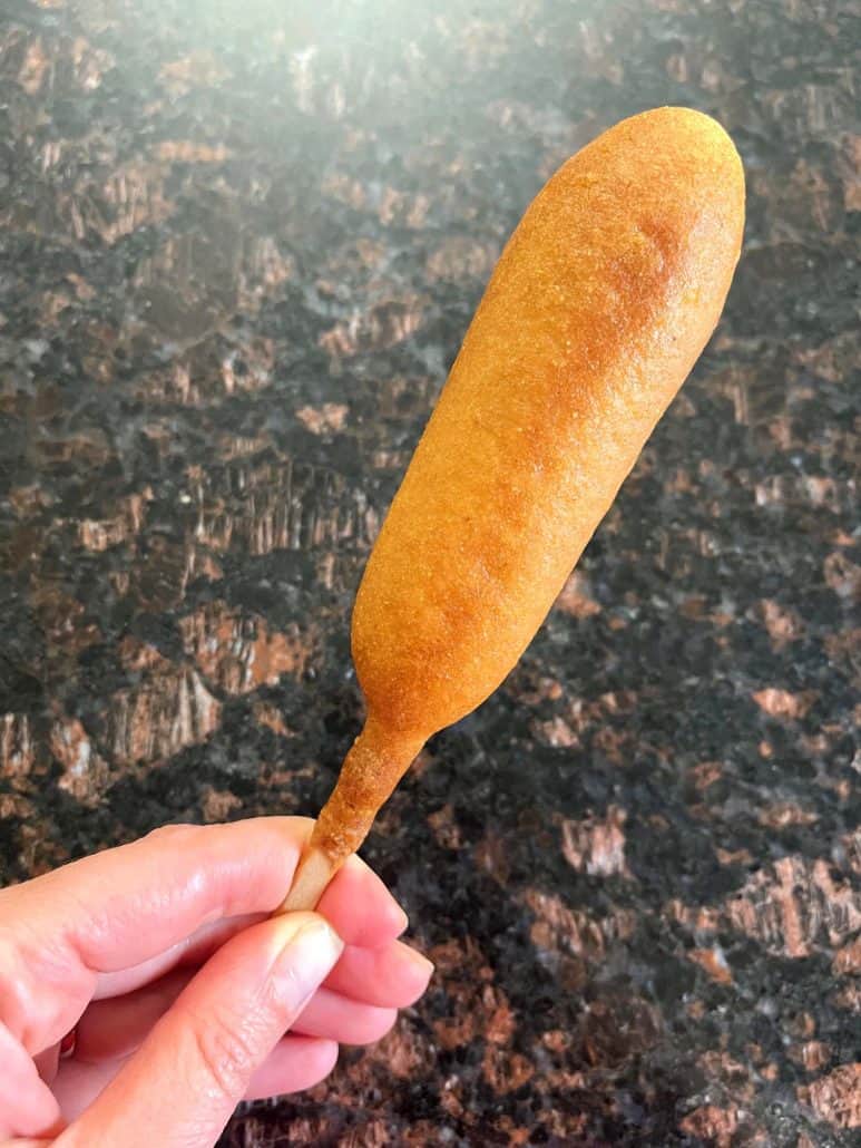 Hand holding a freshly cooked vegan corn dog with a golden, crispy coating.