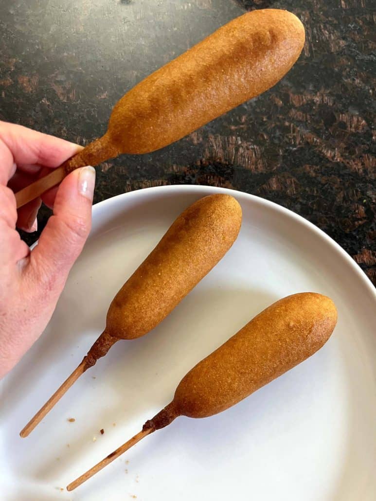 Hand holding one vegan corn dog above a plate, all freshly cooked and golden brown.