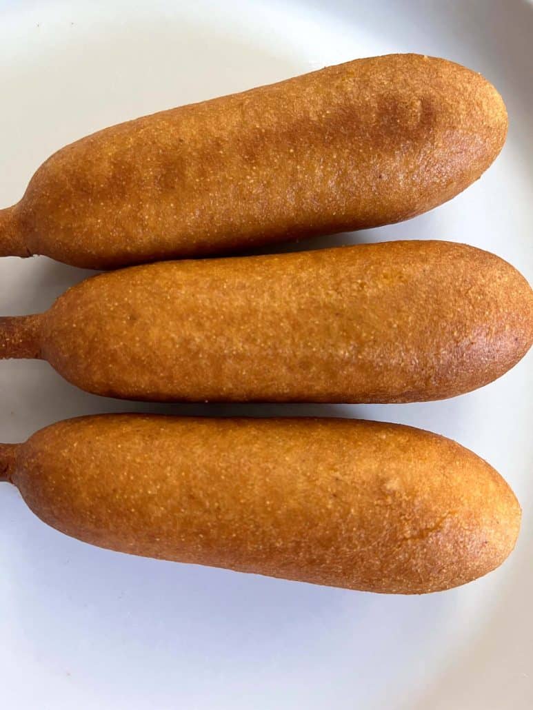 Top view of three golden-brown vegan corn dogs on a white plate, highlighting their crispy coating.