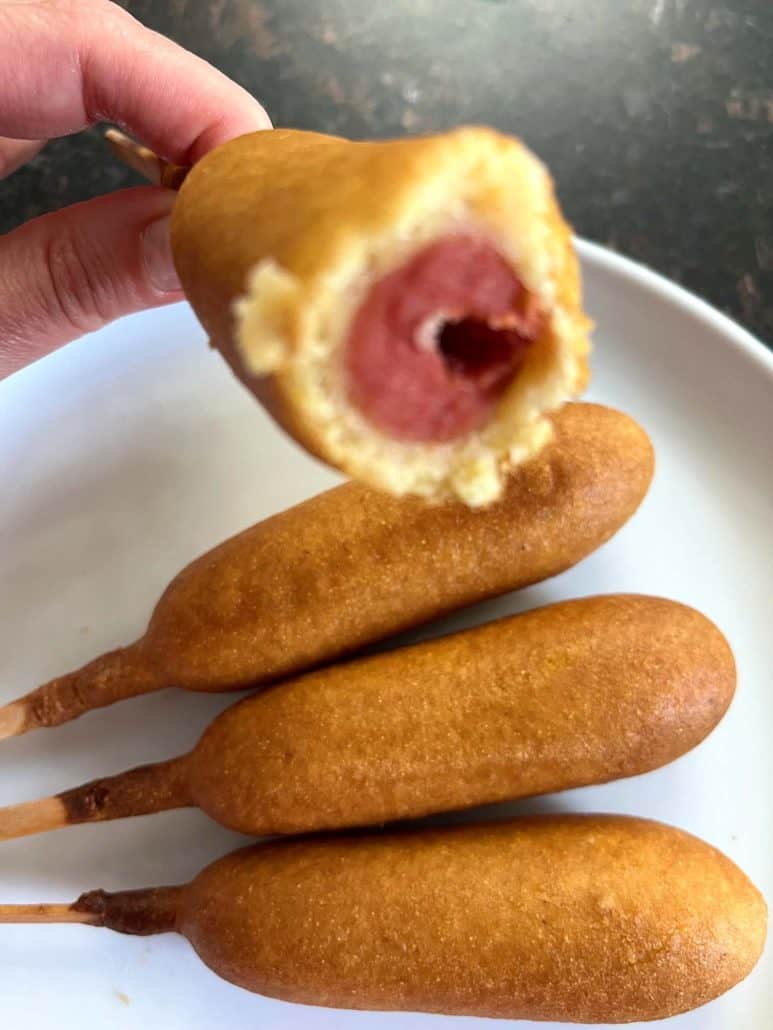 Hand holding a bitten crispy golden vegan corn dog showing the hot dog inside.