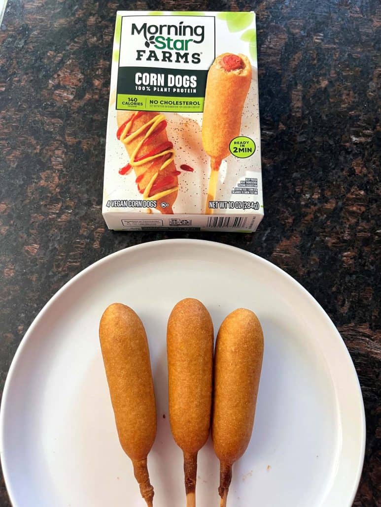 Three cooked vegan corn dogs on a white plate with the MorningStar Farms corn dog package in the background.