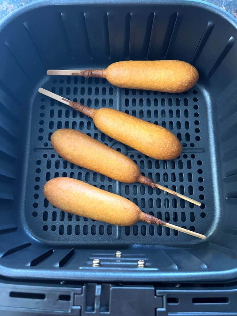 Four vegan corn dogs evenly spaced in an air fryer basket after cooking, showing a crispy golden finish.