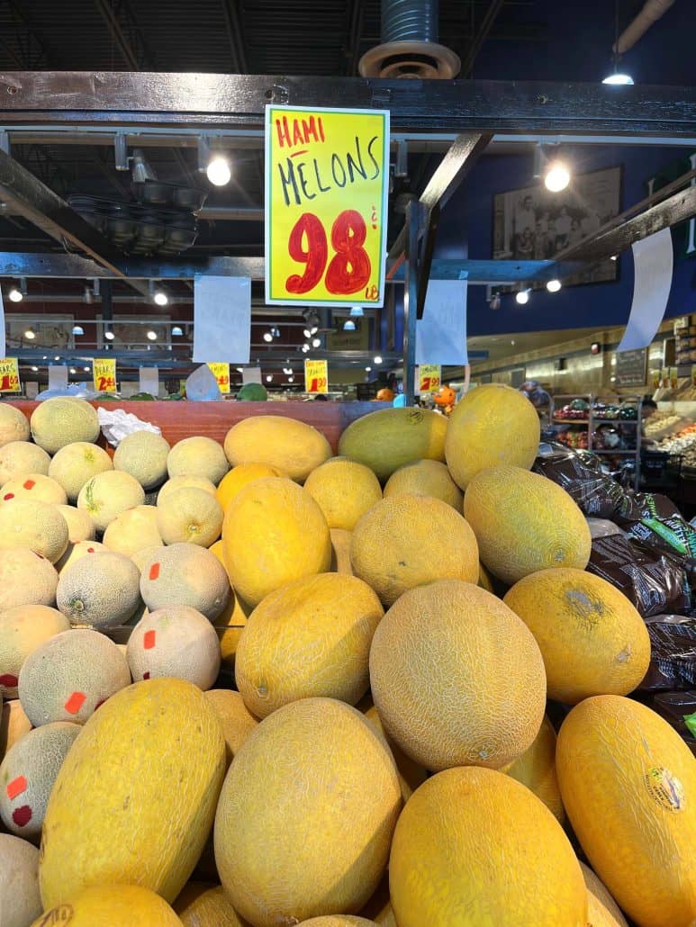 A display of Hami melons at a grocery store, with a handwritten sign above showing the price as 98 cents per pound. The melons, with their yellowish rinds and netted texture, are piled high alongside other types of melons.