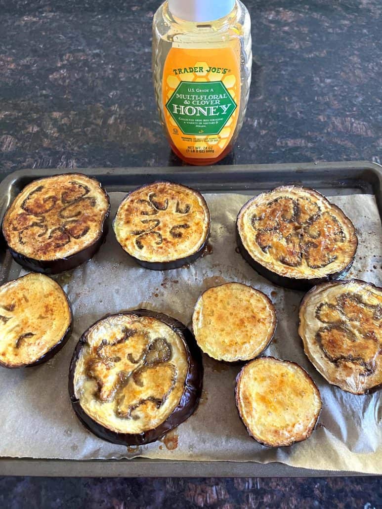 Roasted eggplant slices on a baking sheet with a bottle of honey in the background.