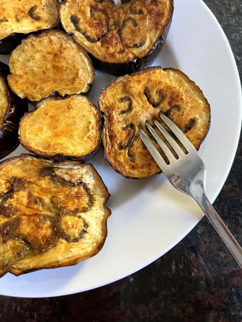 A fork pressing into a golden-brown roasted eggplant slice on a plate.