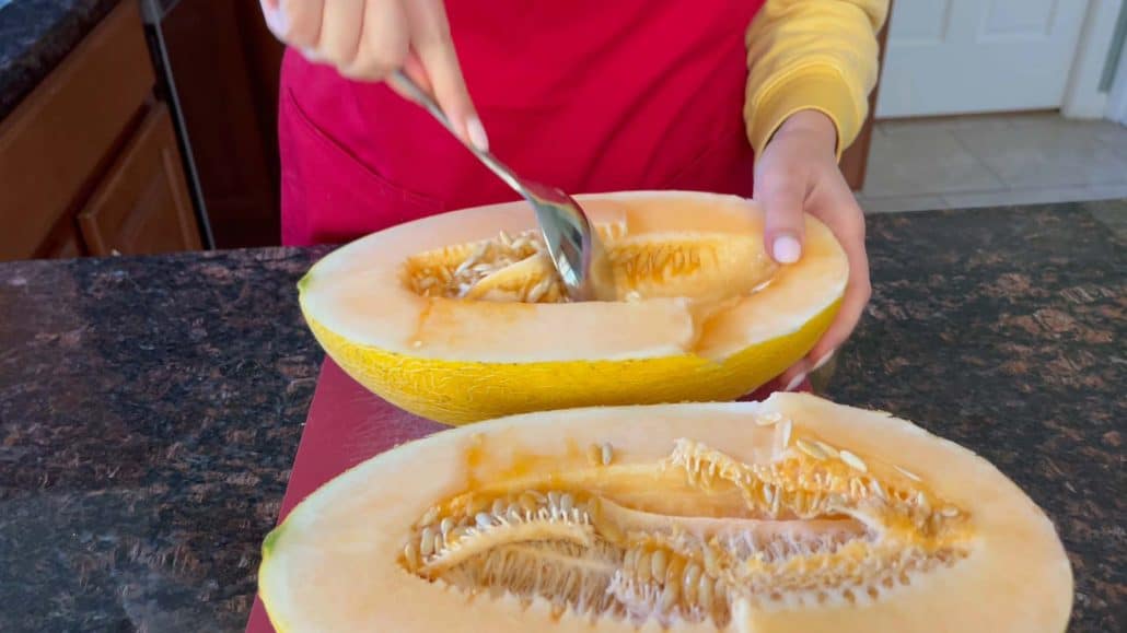 Melanie Mendelson from melaniecooks.com is seen scooping out the seeds from a halved Hami melon in her kitchen. She is wearing a red apron, using a spoon to remove the seeds from the melon’s pale orange flesh, preparing it on a granite countertop.