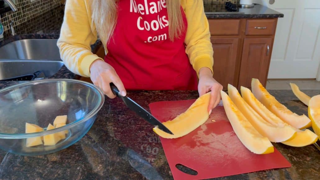Melanie Mendelson from MelanieCooks.com is peeling the rind off Hami melon wedges using a knife on a red cutting board. She is wearing a yellow sweater and a red apron, with some melon cubes already placed in a glass bowl on the countertop. Several peeled melon wedges are visible beside her as she continues preparing the fruit.