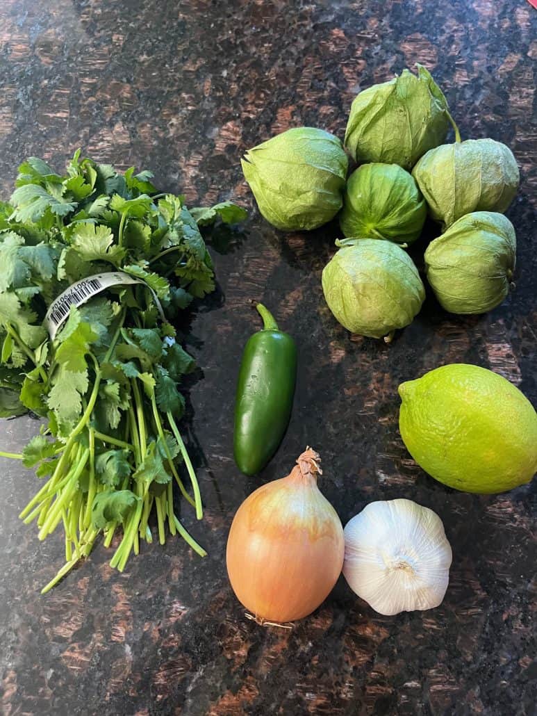 Fresh ingredients for raw tomatillo salsa verde laid out on a countertop, including cilantro, tomatillos, jalapeño, lime, onion, and garlic.