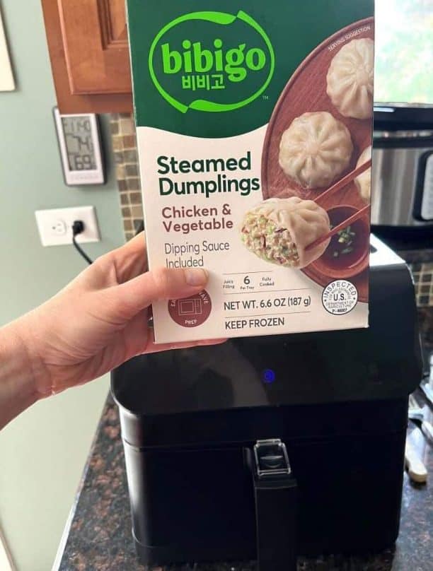 Hand of Melanie Mendelson from melaniecooks.com holding a package of Bibigo Steamed Dumplings in front of an air fryer, ready to be cooked directly from frozen.