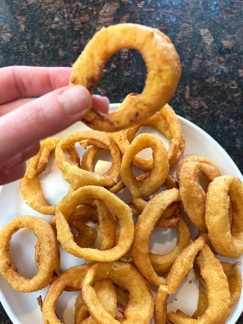 Hand of Melanie Mendelson of melaniecooks.com holding a perfectly crispy golden onion ring above a plate of freshly air-fried onion rings.