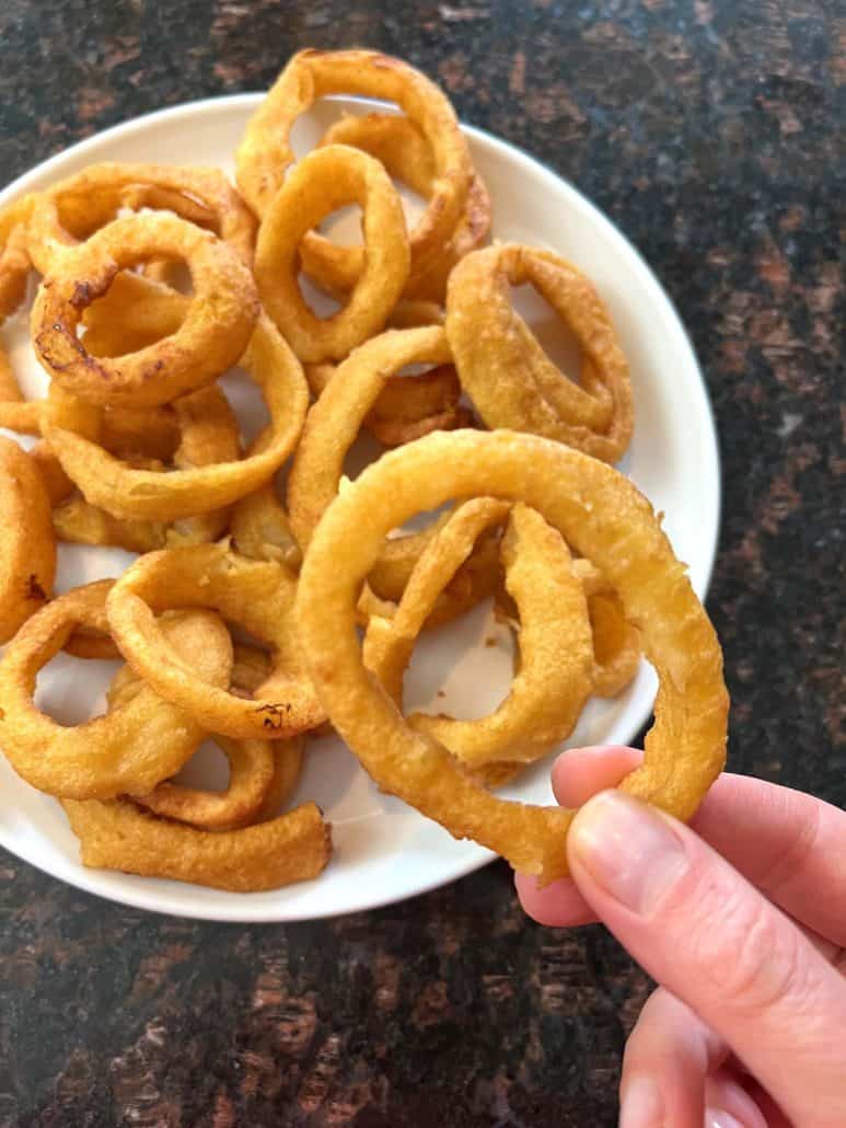 Hand holding a crispy golden onion ring above a plate of freshly cooked onion rings.