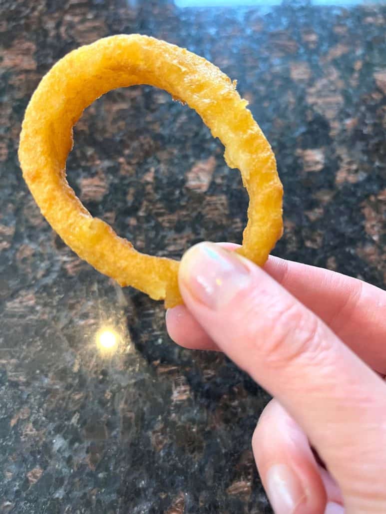 Hand of Melanie Mendelson of melaniecooks.com holding a single crispy, golden onion ring, highlighting its perfect circular shape and crunchy texture.