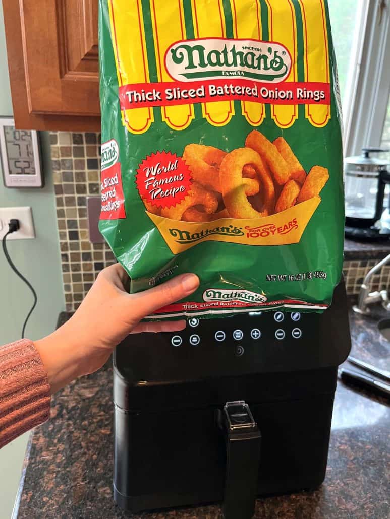 Hand holding a bag of Nathan’s Thick Sliced Battered Onion Rings in front of an air fryer, ready to make a crispy snack.