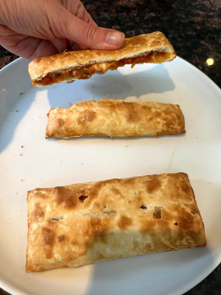 The hand of Melanie Mendelson from melaniecooks.com holding a broken pizza stick to reveal the melted filling inside, with two more crispy pizza sticks on a white plate below.